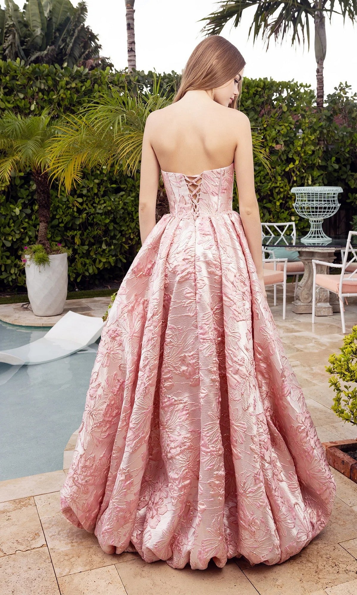 A woman in the Portia and Scarlett XO180 long formal dress, featuring a strapless a-line silhouette with a boned corset bodice and pink textured fabric, stands poolside with her back to the camera, surrounded by greenery and outdoor furniture.